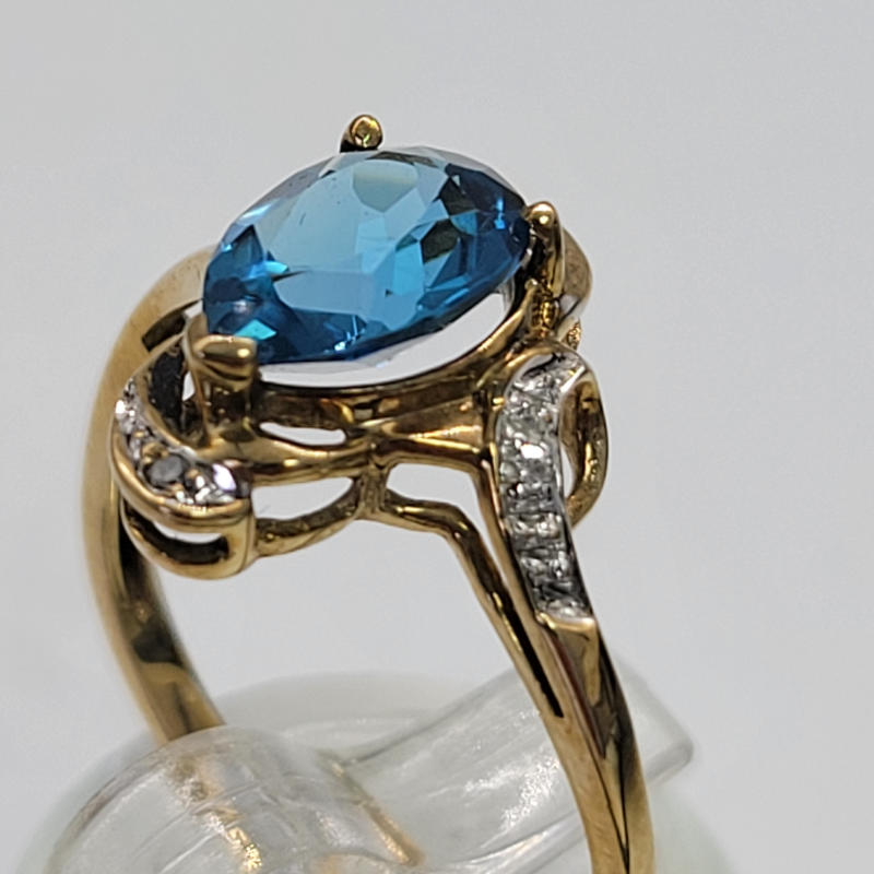 9ct Teardrop Blue Topaz and Diamond Ring An unusual pear-shaped blue topaz, boasting an approximate weight of 1K. This magnificent gemstone, with its captivating hue reminiscent of the deepest oceans, is elegantly nestled within a resplendent 9ct yellow gold setting with a smattering of sparkly diamonds. Every stone in this exquisite ring has been meticulously secured in place with delicate claw settings, ensuring that each facet catches the light in the most enchanting manner. The result is a mesmerizing display of brilliance and sparkle that is sure to captivate all who lay their eyes upon it. Below the topaz is a space in the setting to allow natural light to shine through it. While this remarkable topaz is virtually flawless, it bears a minuscule imperfection in the form of a tiny chip at the bottom of its pointed end. However, this blemish is so inconspicuous that it remains virtually invisible to the unaided eye. Rest assured, the overall beauty and allure of this remarkable gemstone remain unmarred by this minute flaw. For those seeking to adorn their finger with this extraordinary piece of artistry, it is important to note that this ring is available in UK Size N or US Size 6½. The inner circumference measures a precise 53.5mm