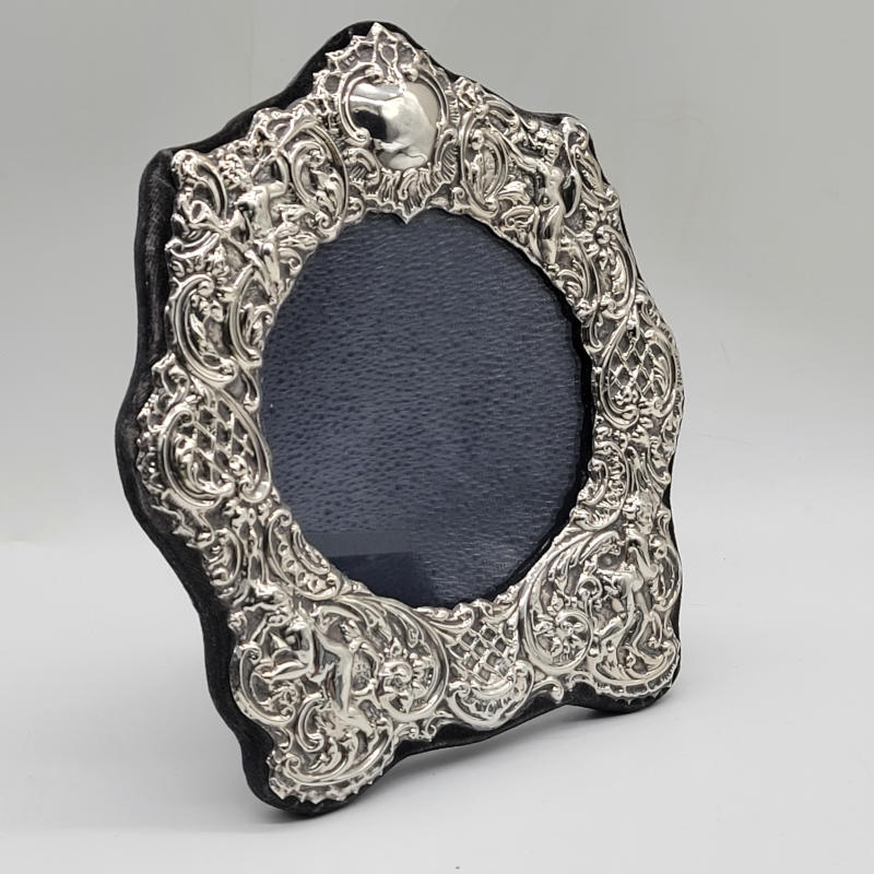 Vintage Silver Cherub Photo Frame An exquisite and meticulously crafted photo frame, adorned with intricate cherub designs, that is sure to captivate your heart. This stunning piece is made of high-quality silver and bears the prestigious London hallmark, guaranteeing its authenticity and craftsmanship. The frame, dating back to the year 1986, holds a timeless charm that will add a touch of elegance to any space. But that's not all! This remarkable frame offers a unique opportunity for personalization. With its empty cartouche, you have the chance to engrave a special message or name, transforming it into a truly one-of-a-kind gift. Imagine the joy on your loved one's face as they receive this personalized treasure, a testament to your thoughtfulness and care. Not only is the front of the frame a work of art, but the reverse side is equally exquisite. Covered in a luxurious dark blue velvet, it adds a touch of sophistication and ensures that your cherished memories are protected with the utmost care. There is also a protective glass front to this wonderful frame. Measuring at a generous 15.5cm in width and 17.5cm in height, this photo frame is the perfect size to showcase your most treasured photographs. The actual space for the photo is circular and measures 9.5cm in diameter. Whether it's a precious family portrait, a cherished moment with friends, or a snapshot of a breath-taking landscape, this frame will beautifully display your memories, allowing you to relive those special moments time and time again.